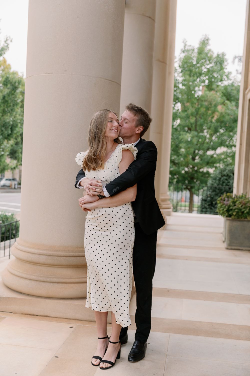 A couple shares romantic kisses in front of elegant stone columns while wearing formal attire.