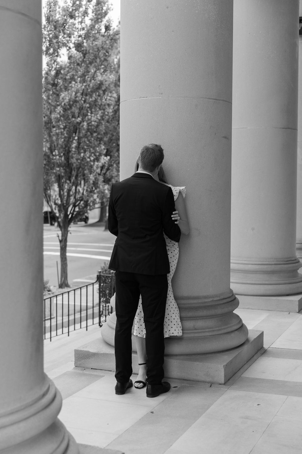 A person in a suit leans against a white classical column in this black and white architectural photograph.