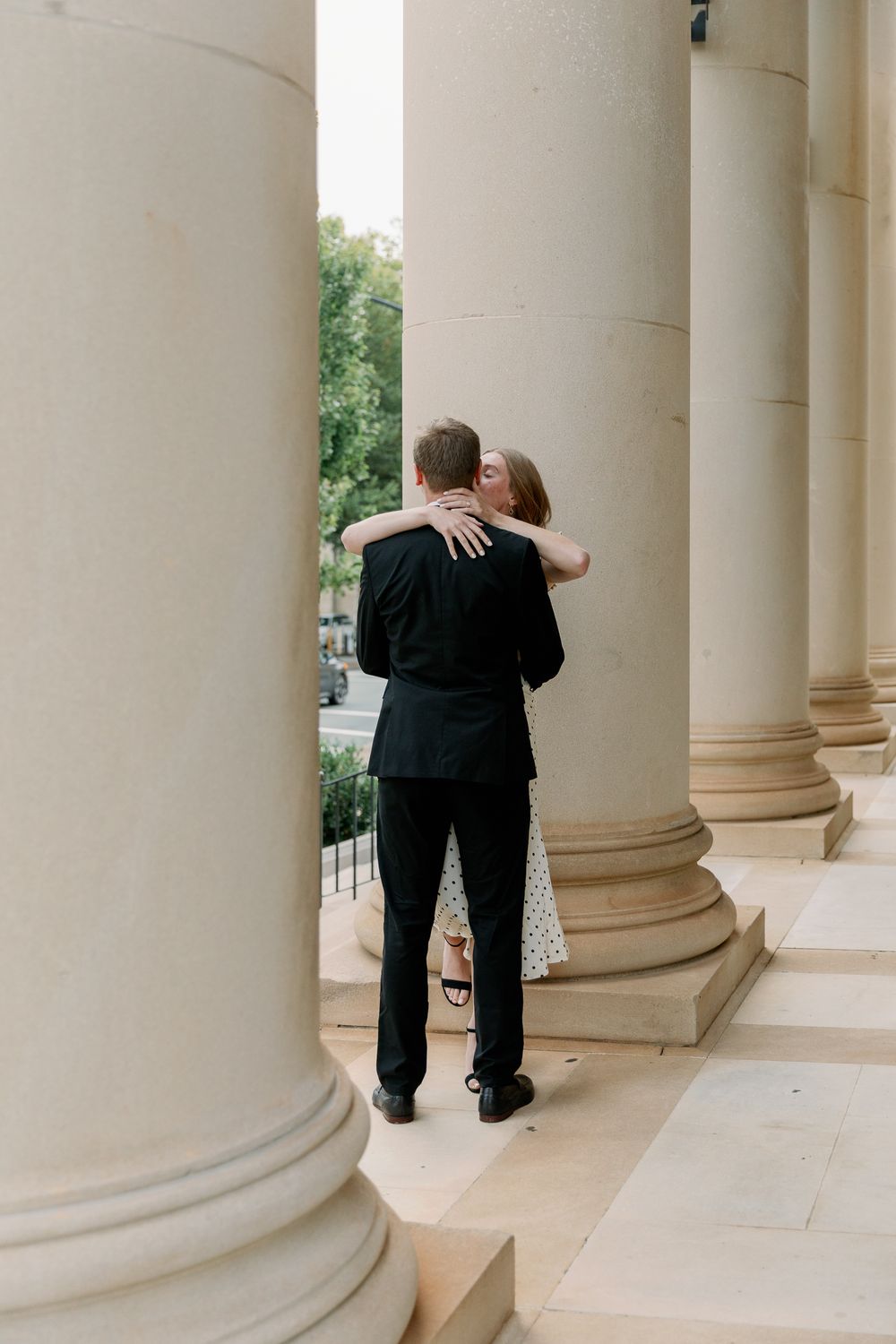 A couple embraces between classical stone columns in an elegant architectural setting.