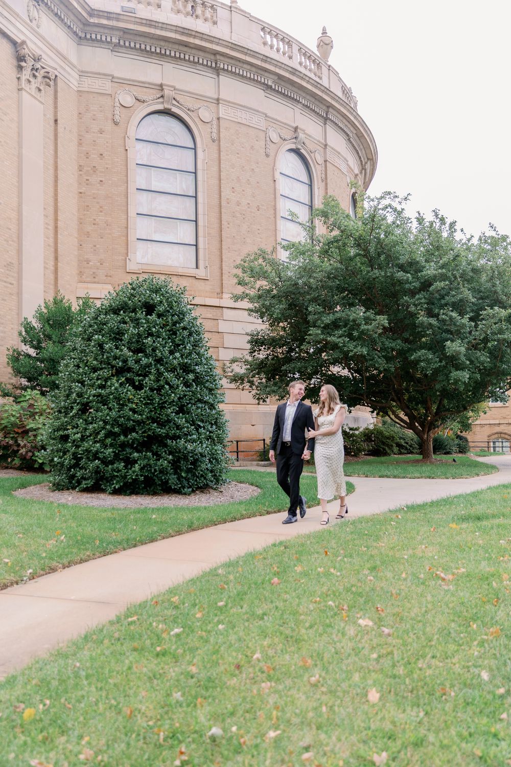 A couple strolls along a curved pathway beside a grand stone building with arched windows and manicured landscaping.