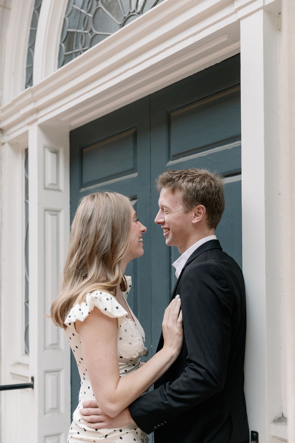 A couple laughs and embraces joyfully in front of a white storefront with blue doors.