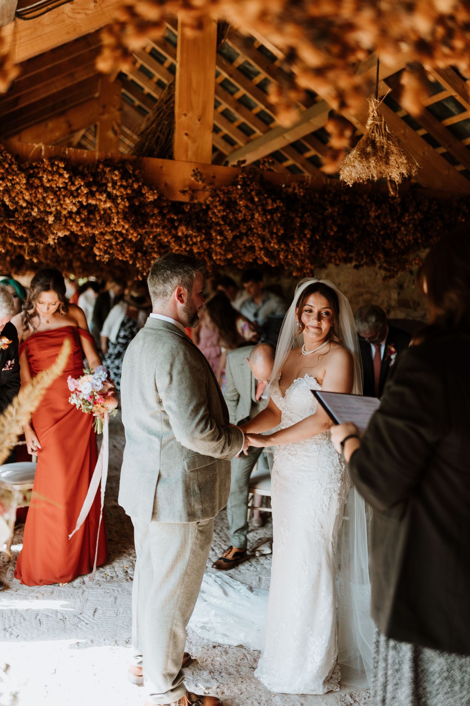 A couple exchanges vows during a rustic barn wedding ceremony under dried flower decorations and warm lighting at Lyde Court, Hereford