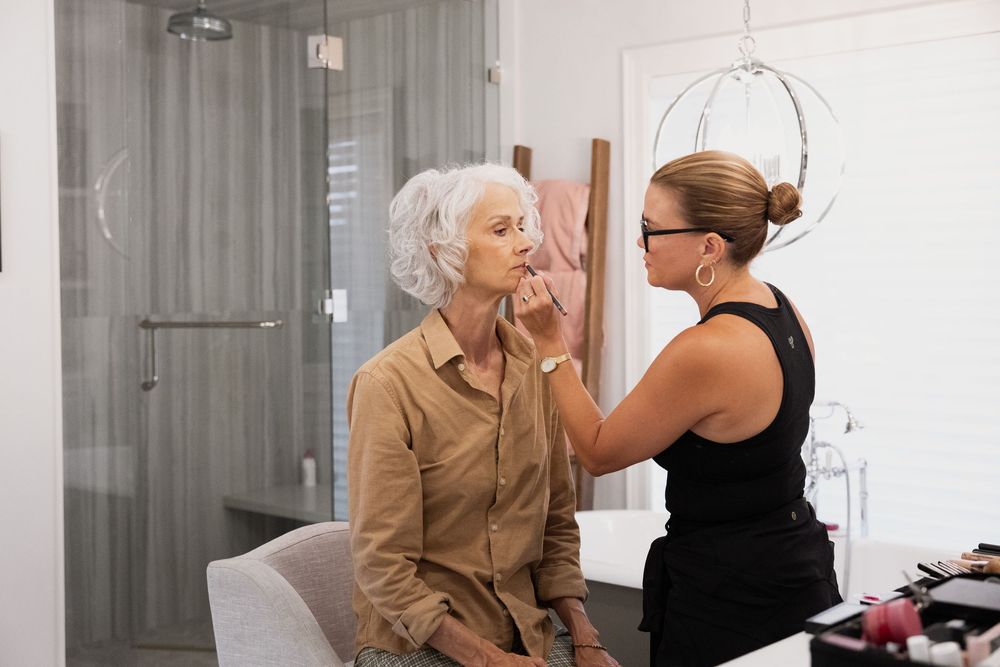 A person in a beige jacket sits while someone in black assists with wedding day preparations.