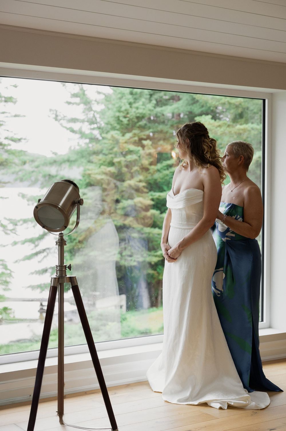 A wedding preparation scene by large windows overlooking pine trees with vintage-style floor lamps.