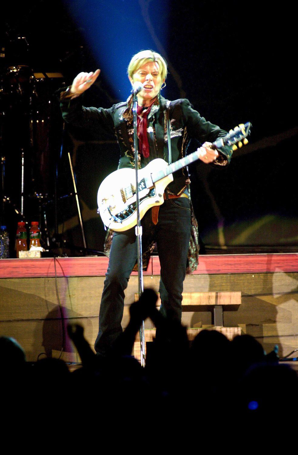 A performer in a black jacket plays a white guitar on stage with dramatic blue lighting during a concert performance.