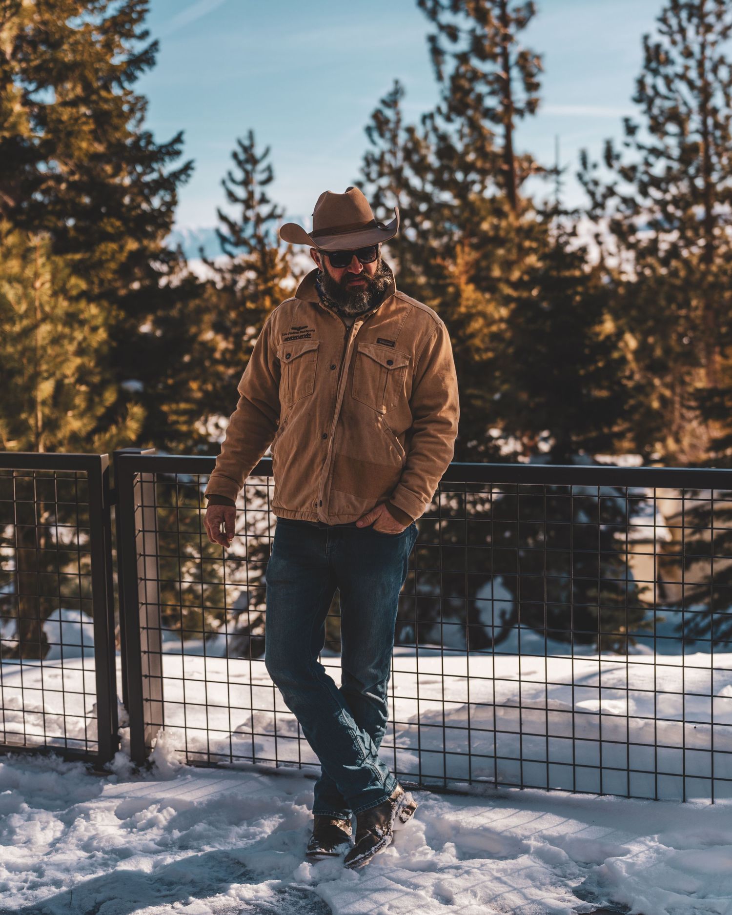 Person in brown jacket and hat poses against snowy winter landscape with evergreen trees in the background.