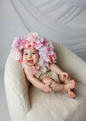 Baby wearing pink floral accessories lies on white fluffy cushion during professional photoshoot.