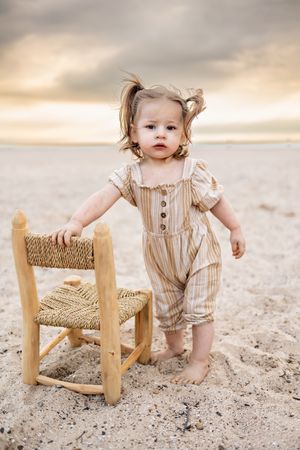 Toddler in striped romper plays with wooden chair on sandy beach during sunset.