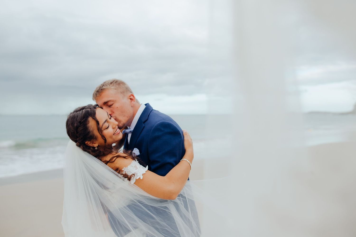 Groom in navy suit lifts and kisses bride wrapped in flowing veil on a cloudy beach.