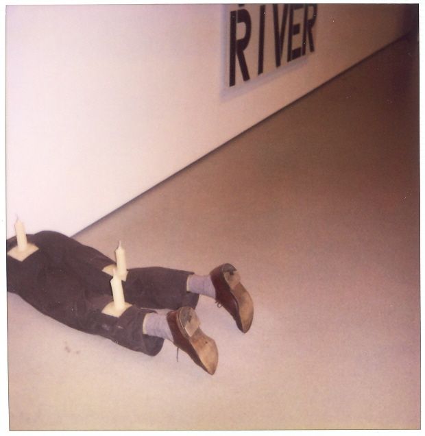 Legs wearing brown shoes and dark pants lying on floor beneath text reading 'RIVER' on a wall in an exhibit at the MoMA in New York City.