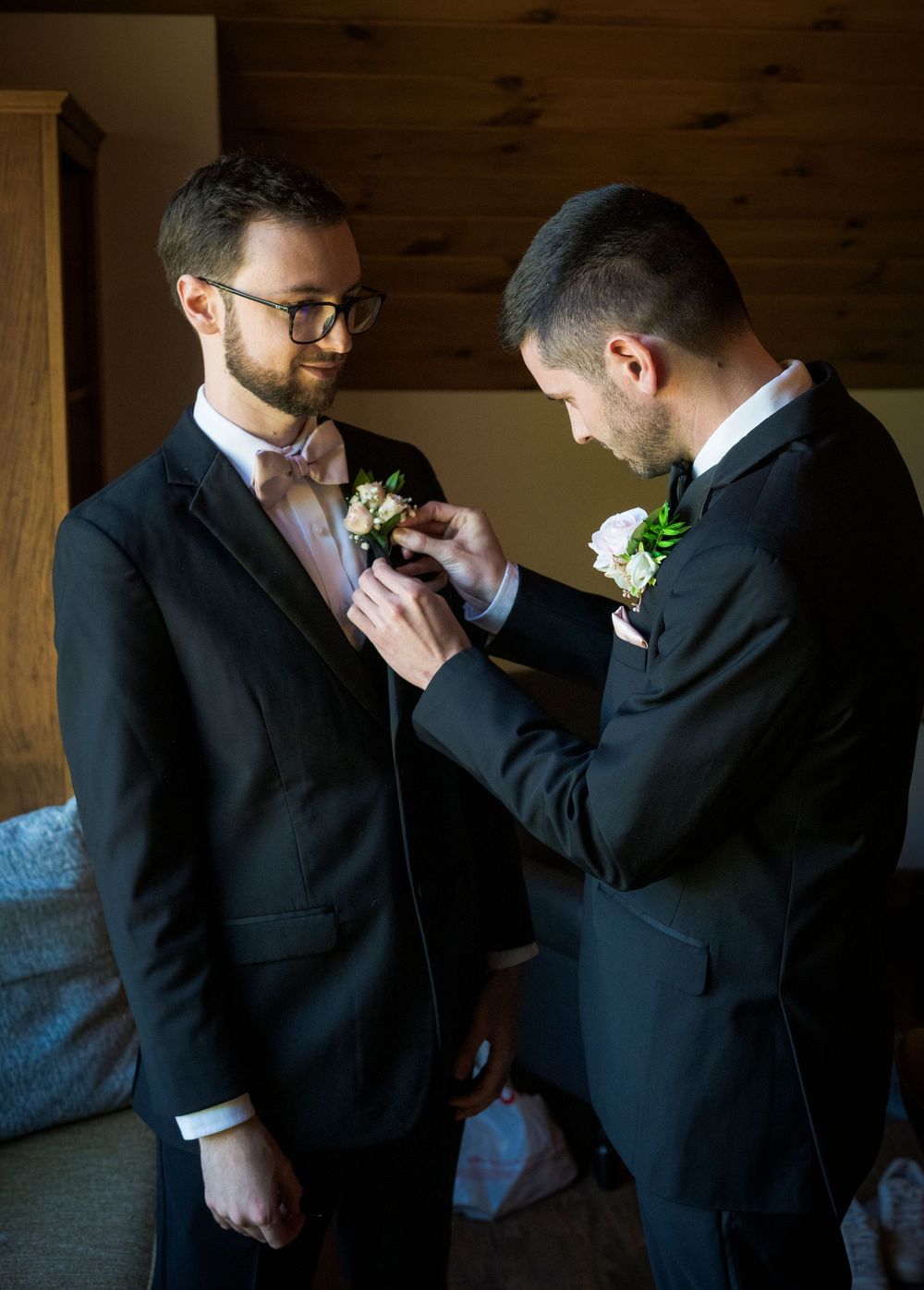 Groomsman pins boutonniere on groom wearing black tuxedo and pink bow tie indoors.