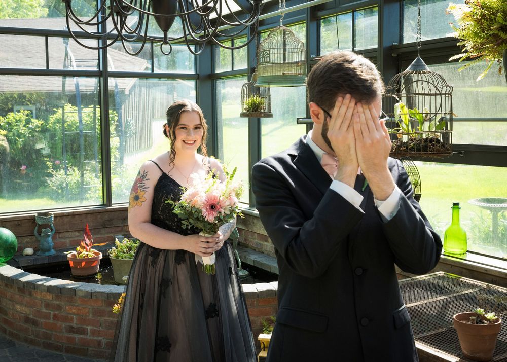 Bride in black gown surprises groom covering his eyes during wedding first look reveal indoors.