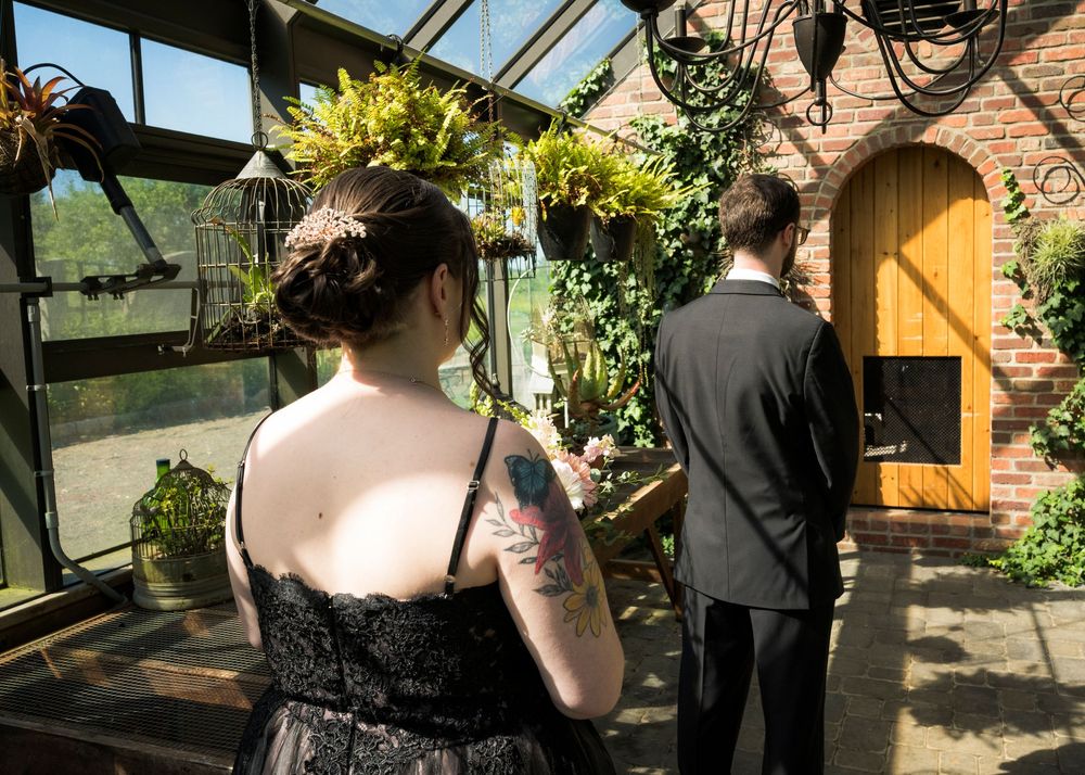 Wedding guest in black dress with floral tattoo and groom in gray suit standing in lush greenhouse venue.