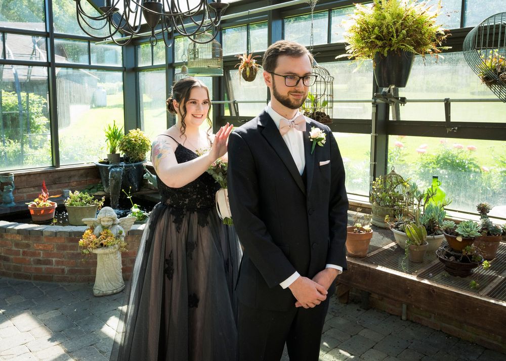 Bride in black wedding gown tapping groom shoulder for first look reveal inside botanical greenhouse wedding venue.