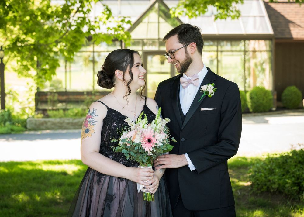 Bride in black floral gown and groom in black tuxedo sharing romantic moment outdoors holding pink flower bouquet.