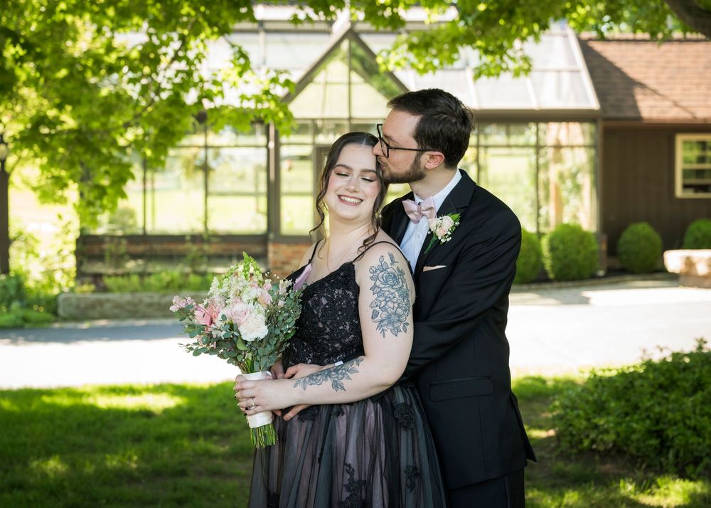 Groom in black tuxedo kisses bride wearing black lace wedding gown while she holds a floral bouquet.