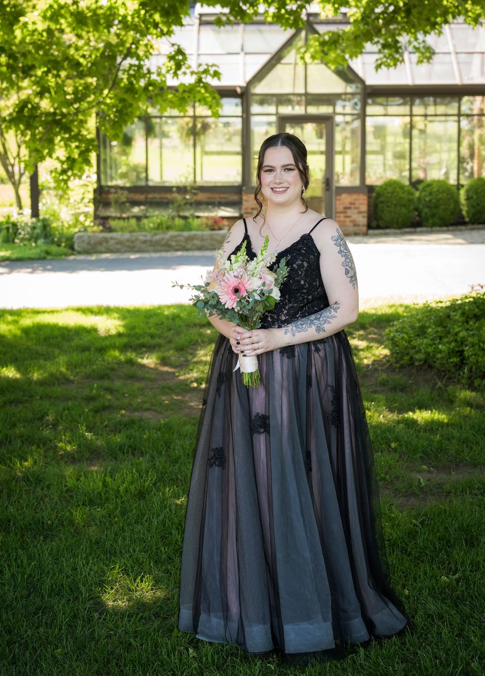 Bride in black lace gown holding pink floral bouquet standing on green lawn with greenhouse backdrop.