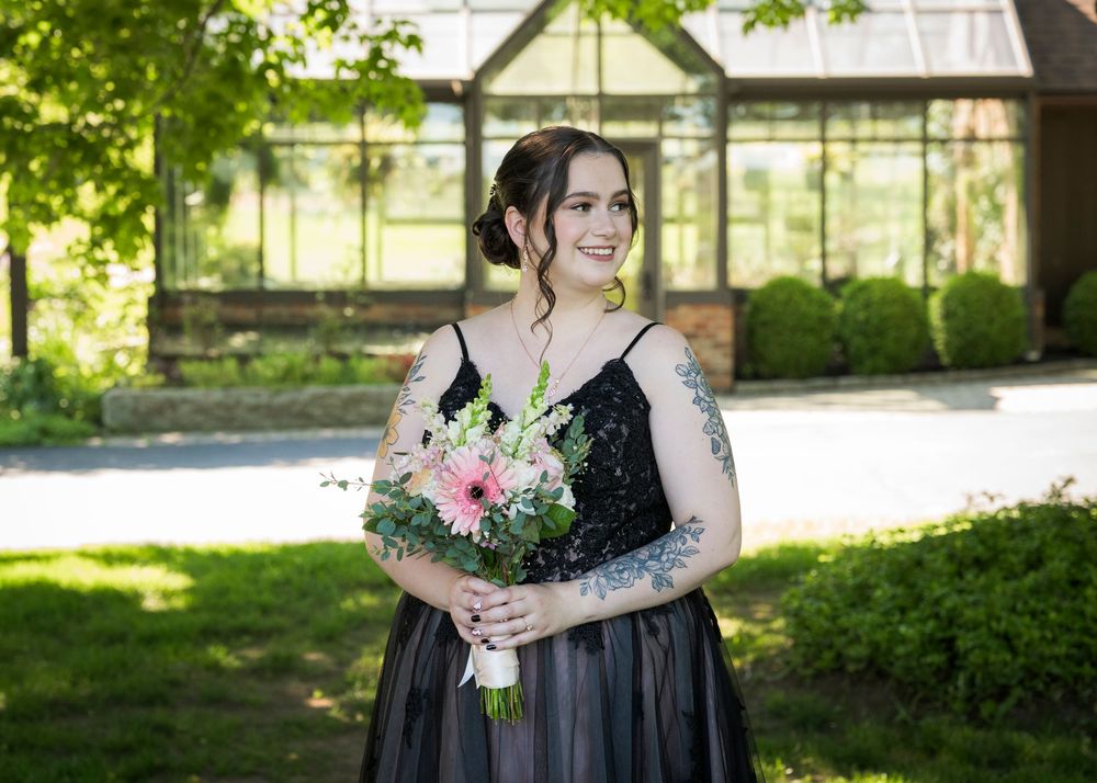 Bridesmaid in black gown holding pink and white floral bouquet standing outdoors in sunlight.