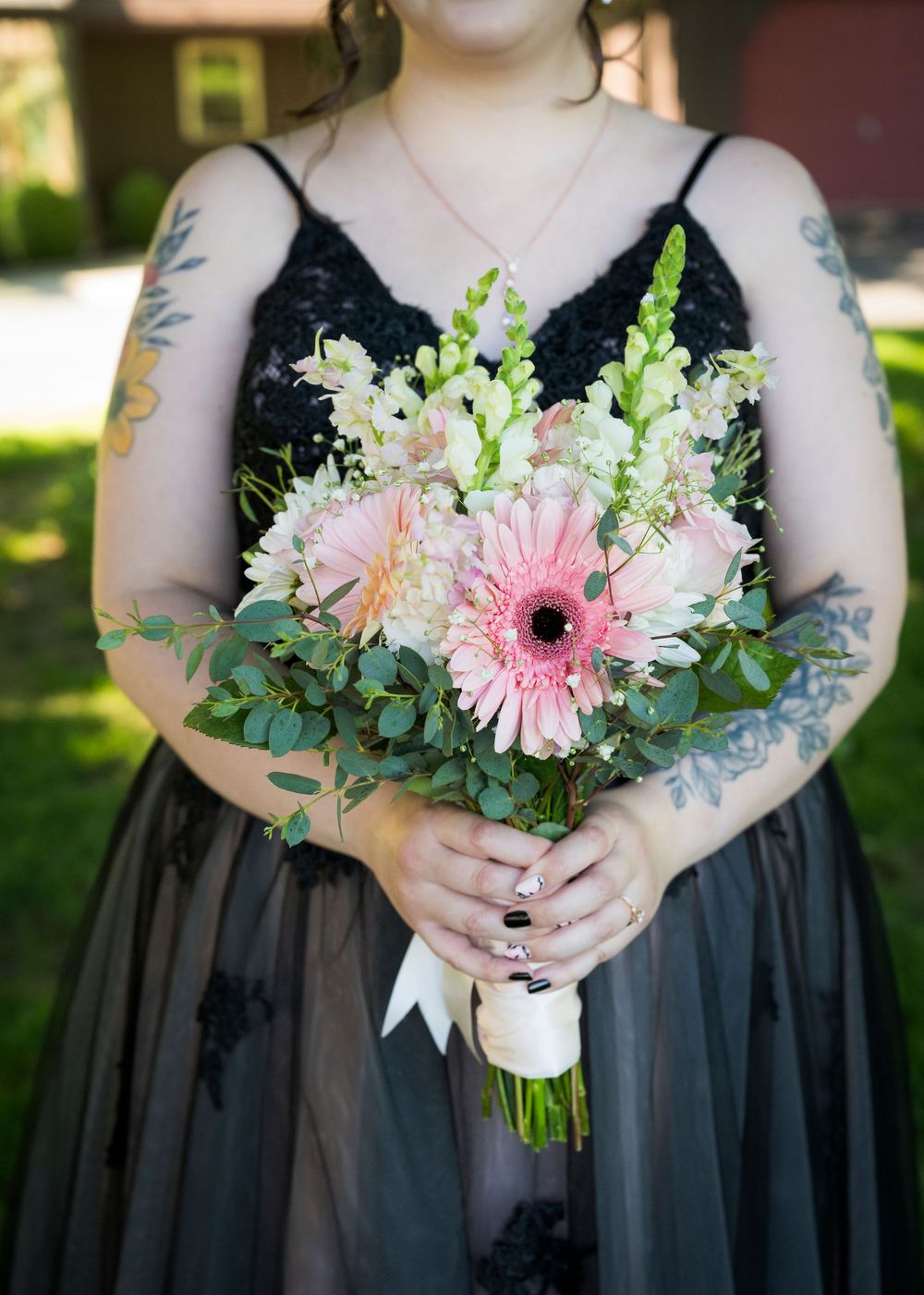 Bride in black lace gown holds pink gerbera daisy and white flower wedding bouquet outdoors.