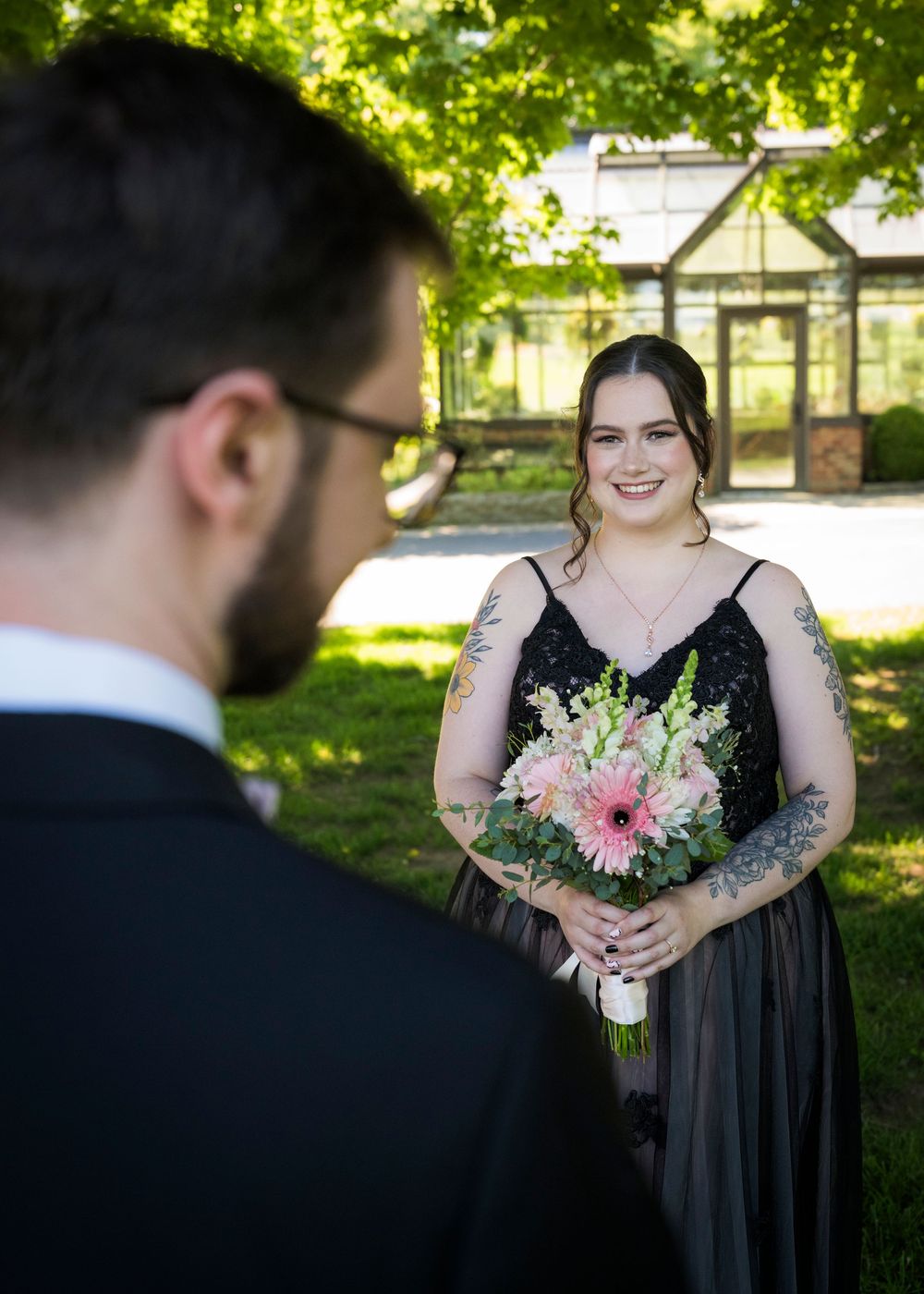 Smiling bridesmaid in black dress holding pink floral bouquet during outdoor wedding ceremony.