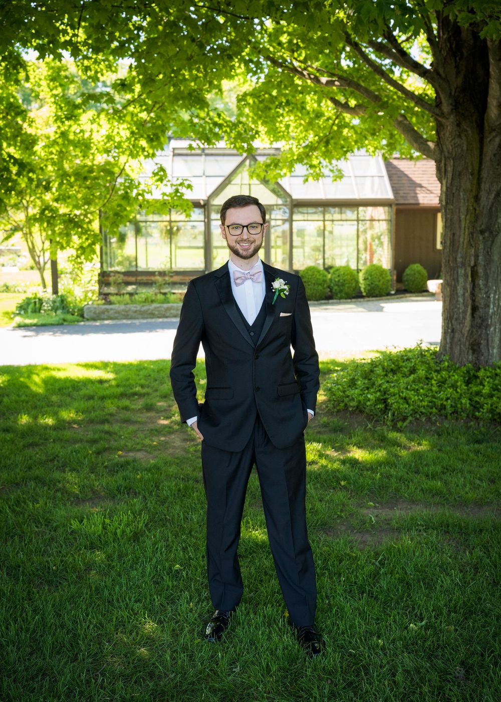 Groom in black suit with pink boutonniere and bow tie standing on green lawn at outdoor venue.