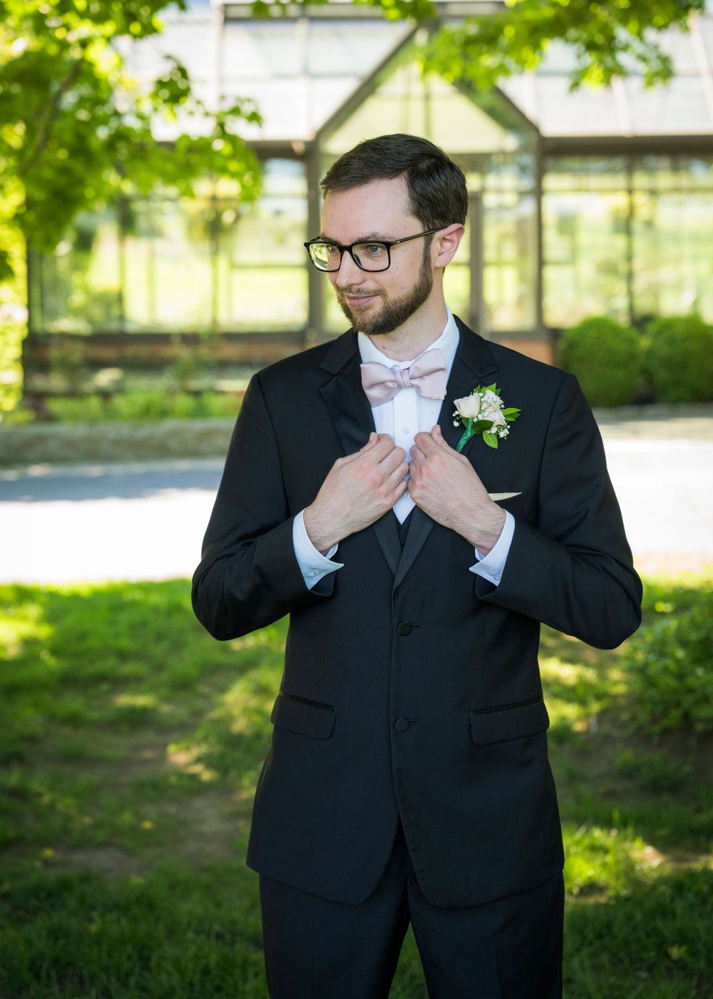 A groom in a black tuxedo with pink bow tie and boutonniere adjusts his jacket outdoors.