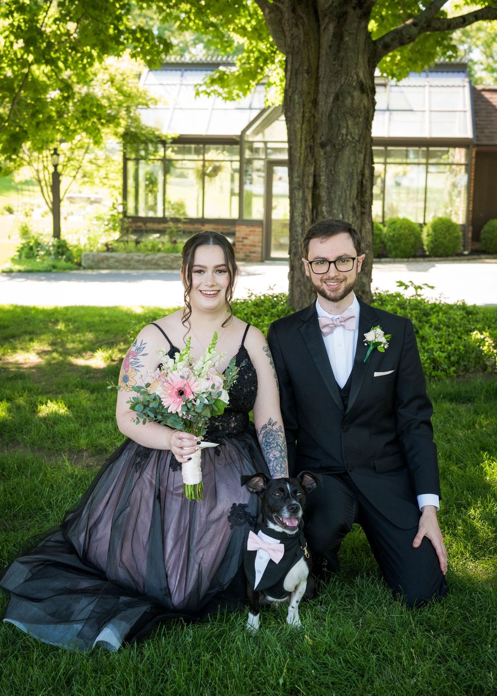 Bride and groom in black wedding attire sit on grass with Boston Terrier dog in pink bow tie.