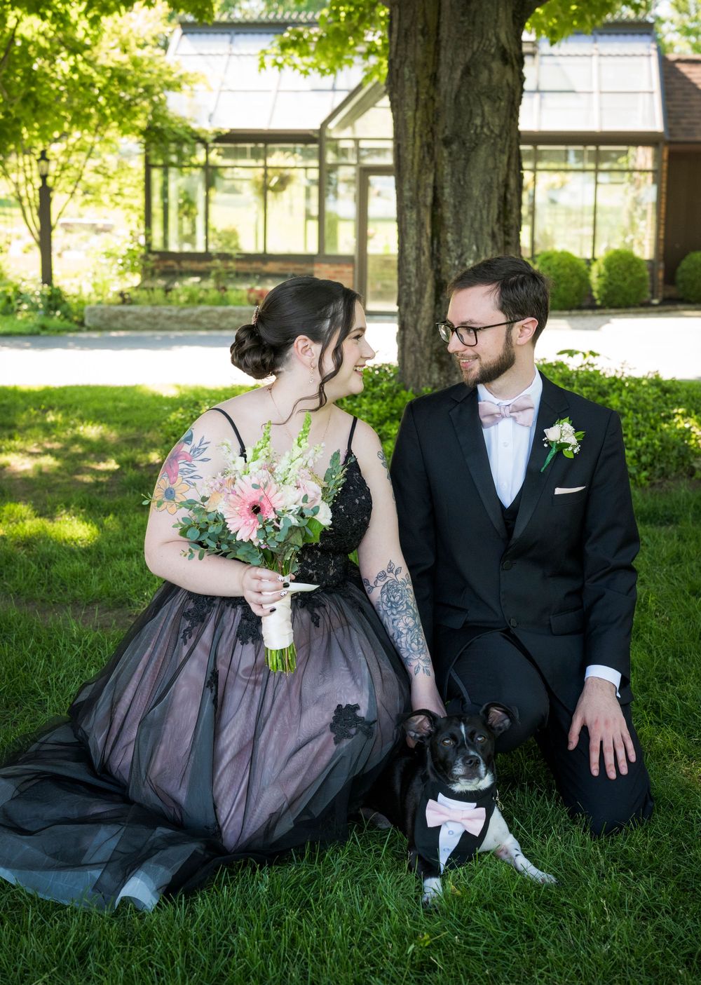 Bride and groom in black wedding attire gaze at each other seated on grass with their dog.