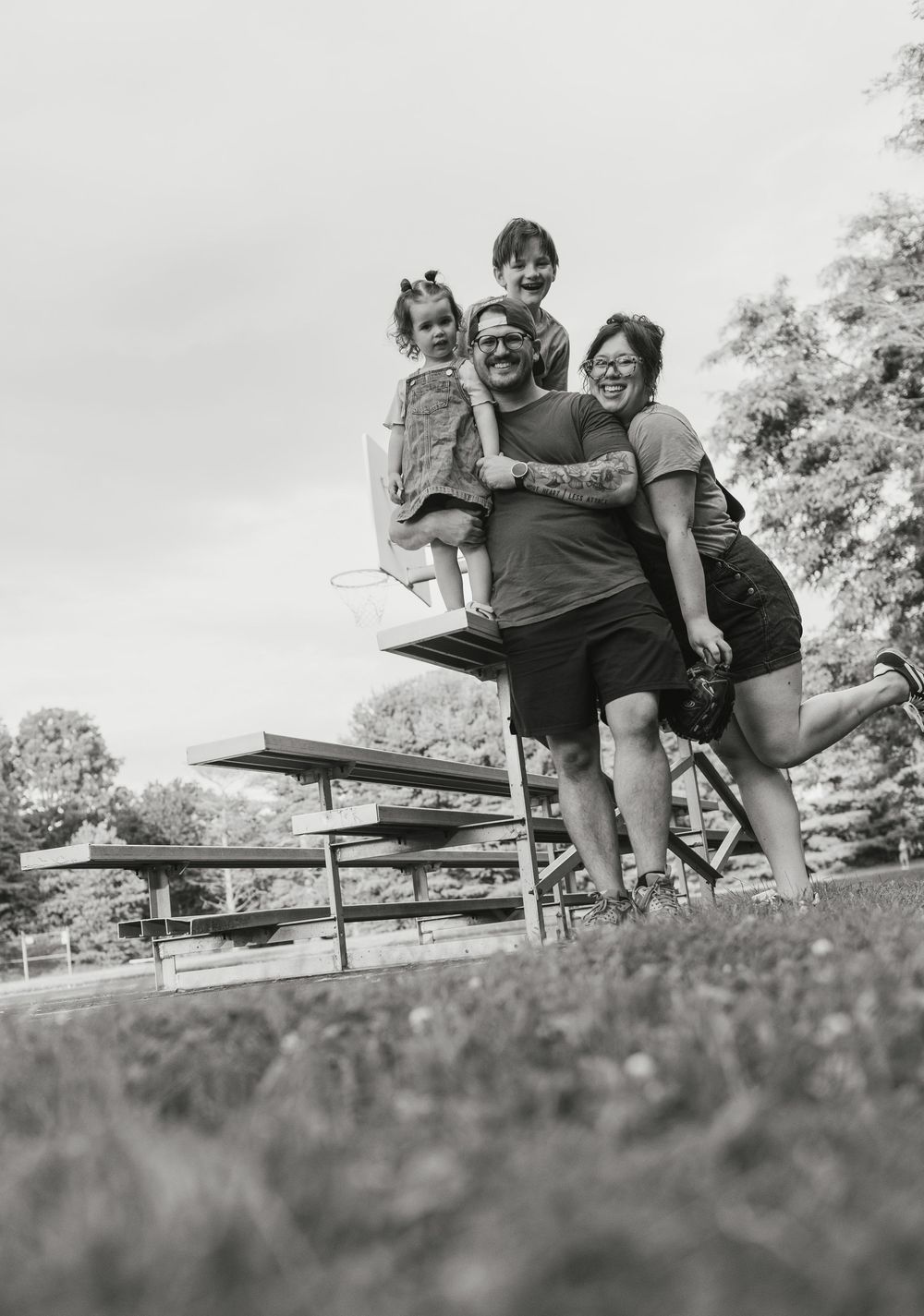 Black and white photo of a joyful family of four posing outdoors near bleachers in a park.