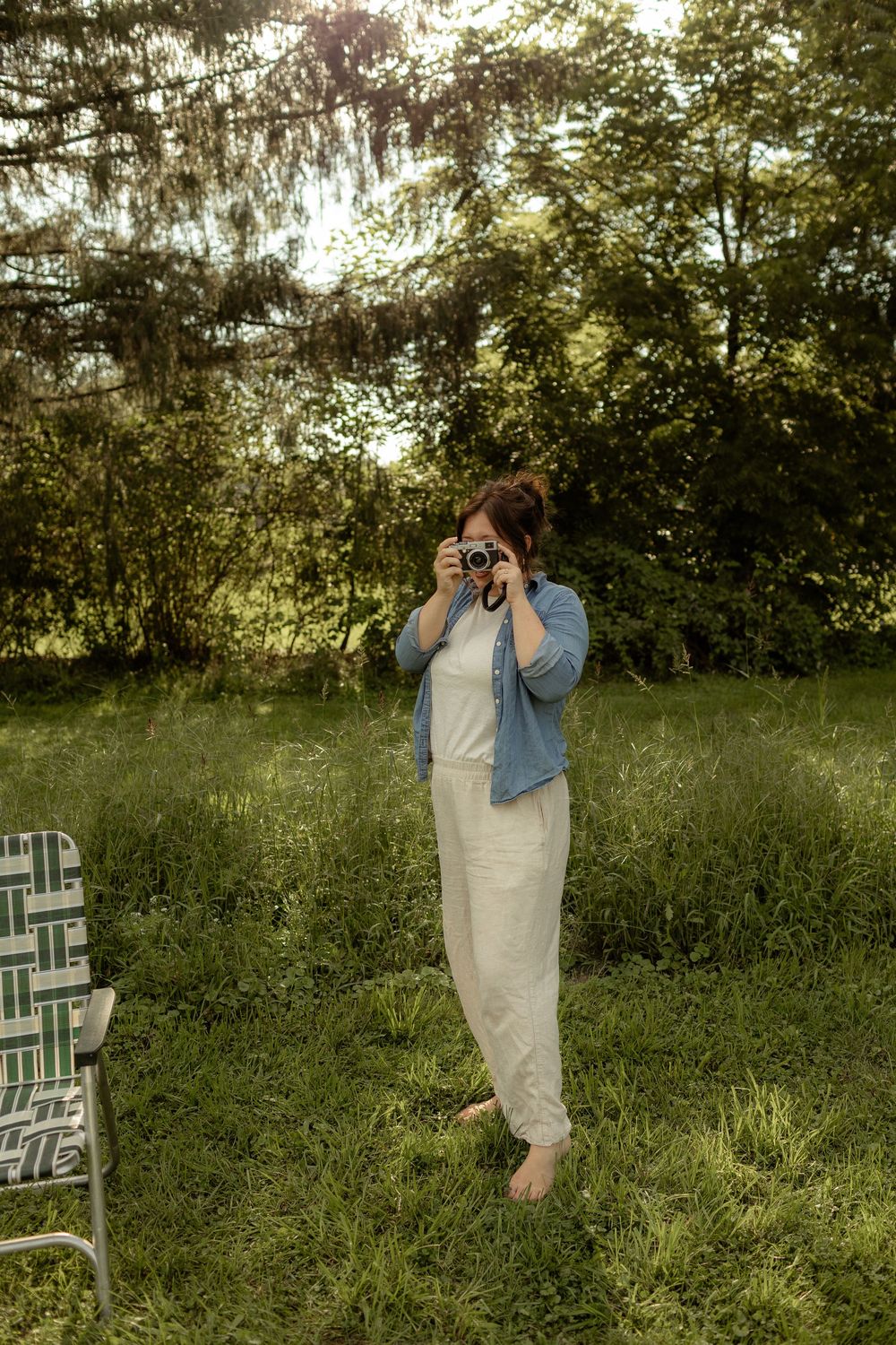 A barefoot woman in white pants and denim shirt photographs outdoors in a lush green garden setting.