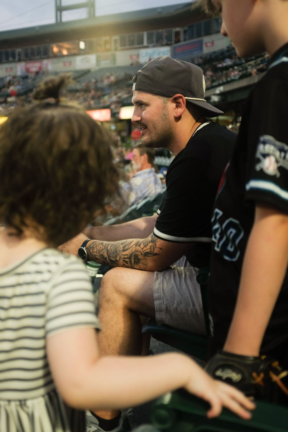 Tattooed baseball player in a cap interacts with young fans in stadium seating during an evening game.