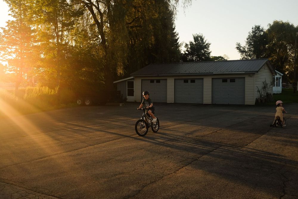 Child riding bike and another playing near garage at golden sunset hour on residential driveway.