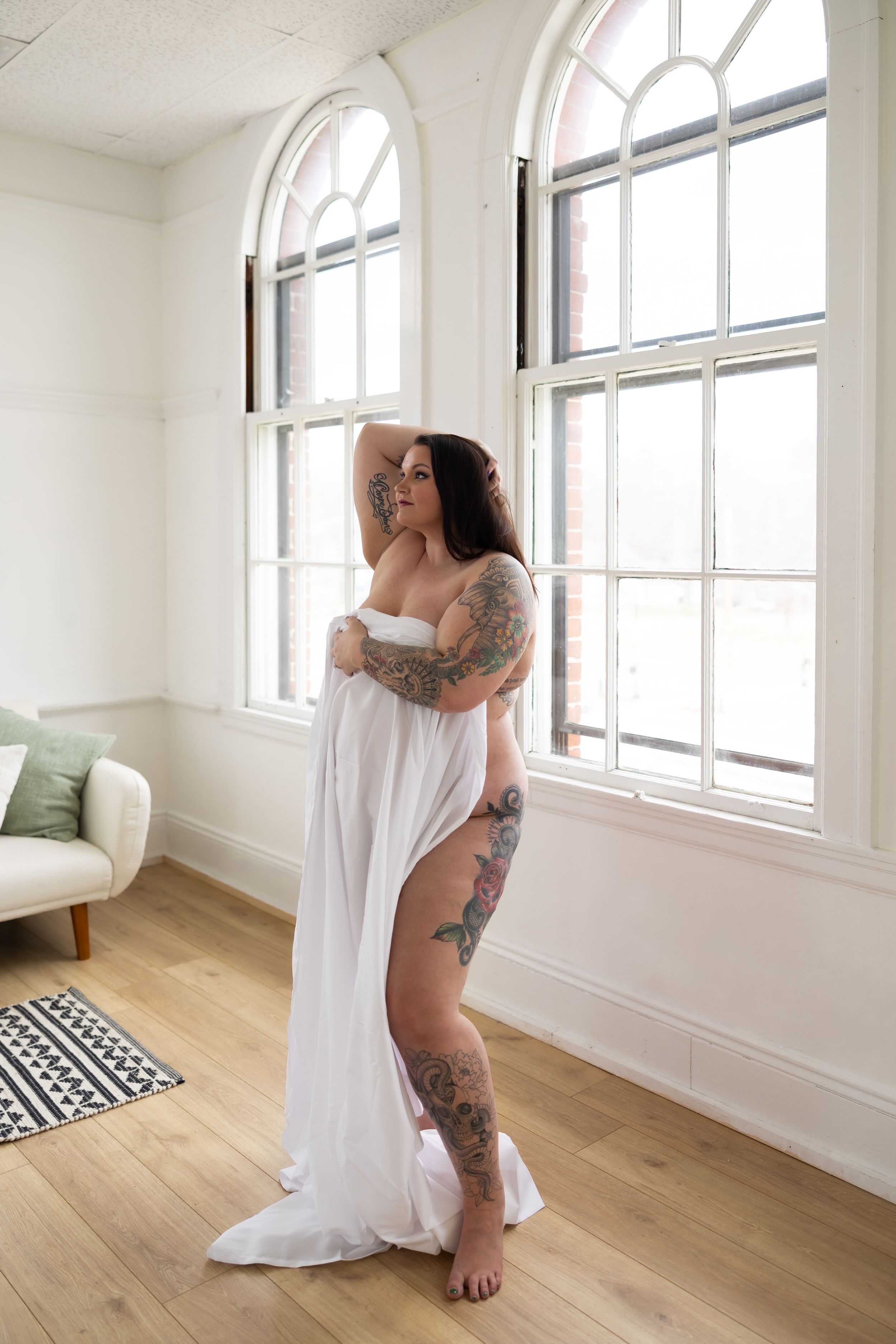 A person in a flowing white fabric poses gracefully by tall arched windows in a bright minimalist room with hardwood floors.