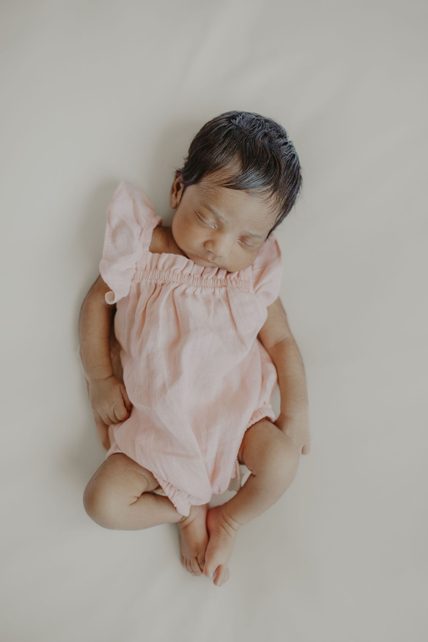 Sleeping newborn baby girl in a pink ruffle romper lying on a cream background.