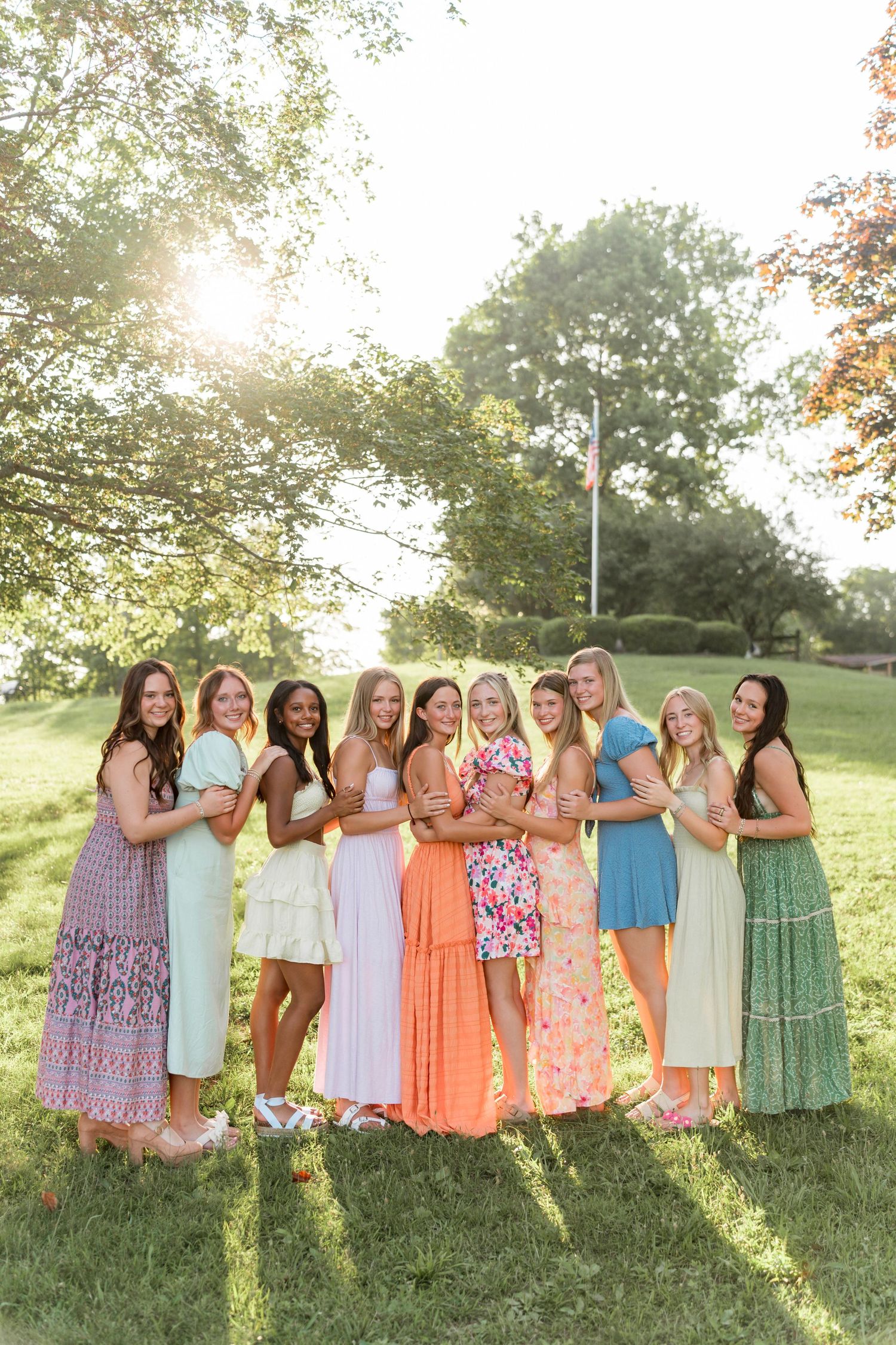 Group of women in pastel summer dresses pose together on grassy hill during golden hour sunset.