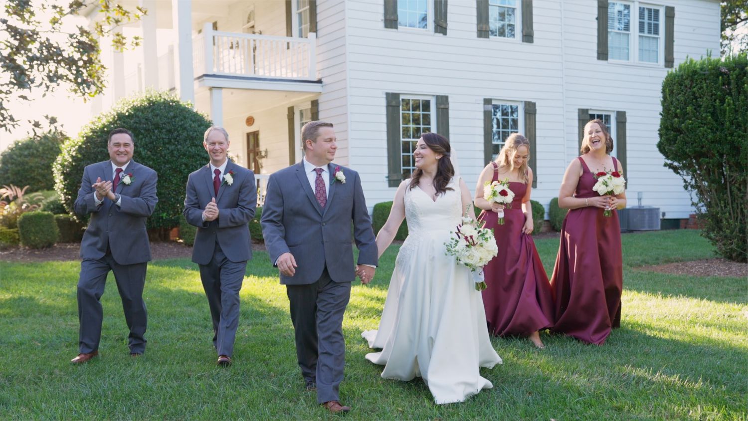 Wedding party members in burgundy dresses and gray suits walk together on a grassy lawn at an outdoor venue.