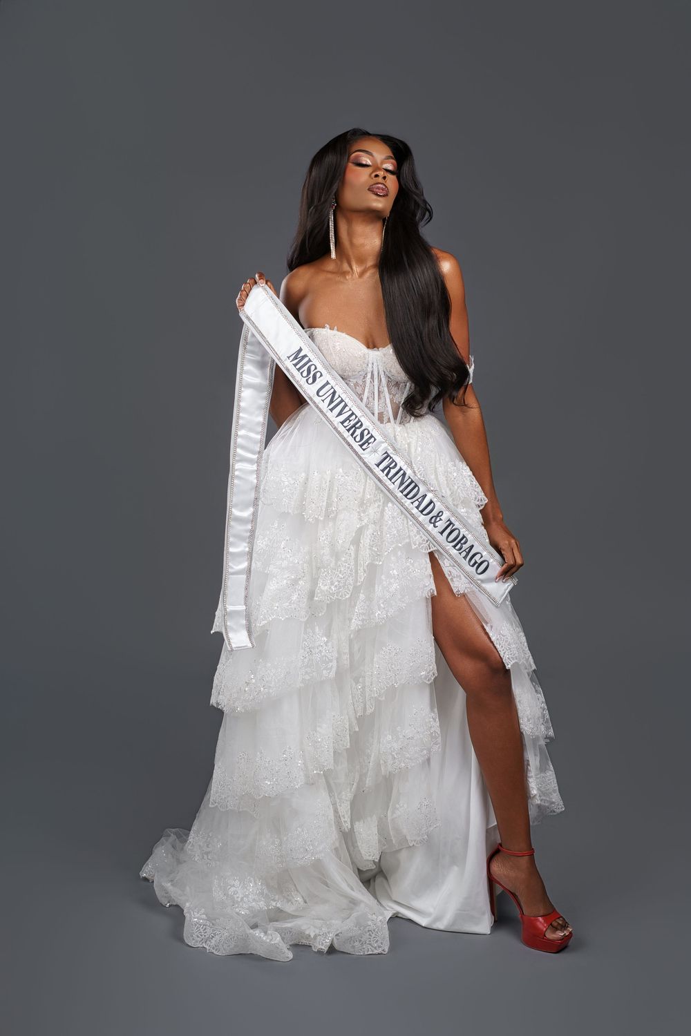 A beauty pageant contestant in a white flowing gown and red heels poses elegantly with a sash against a gray backdrop.