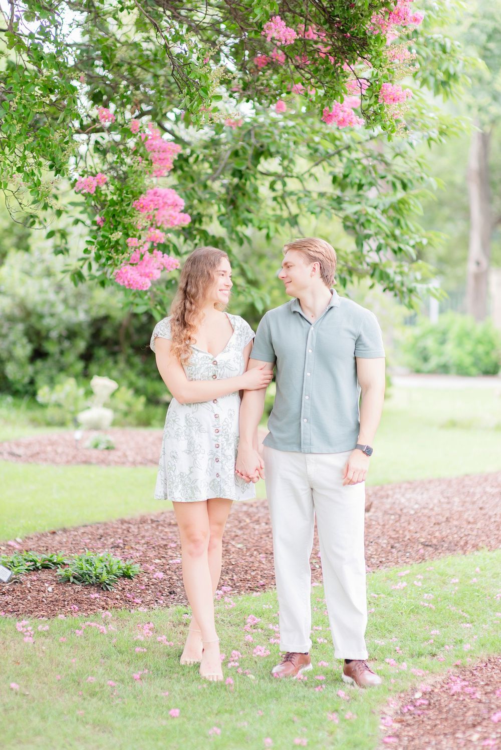 A couple shares a romantic moment under blooming pink crepe myrtle trees in a lush garden setting.