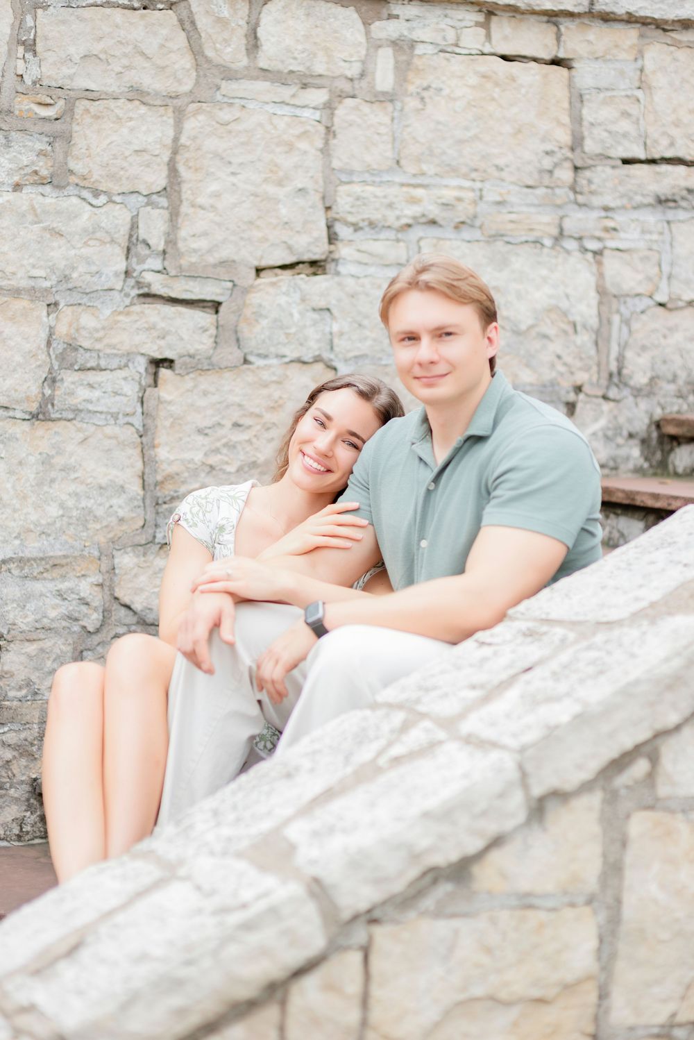 A couple sits together on stone steps against a rustic wall in a romantic outdoor setting.