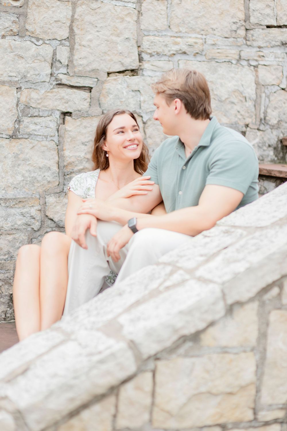 A romantic couple shares a joyful moment while sitting together against a rustic stone wall backdrop.