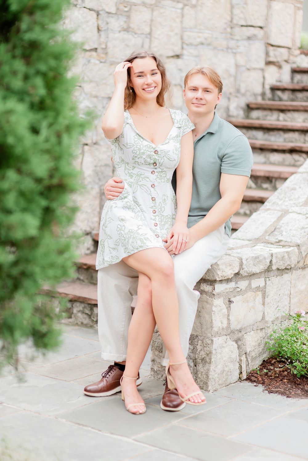 A romantic couple sits on stone steps, she in a floral dress and he in a sage polo and white pants.