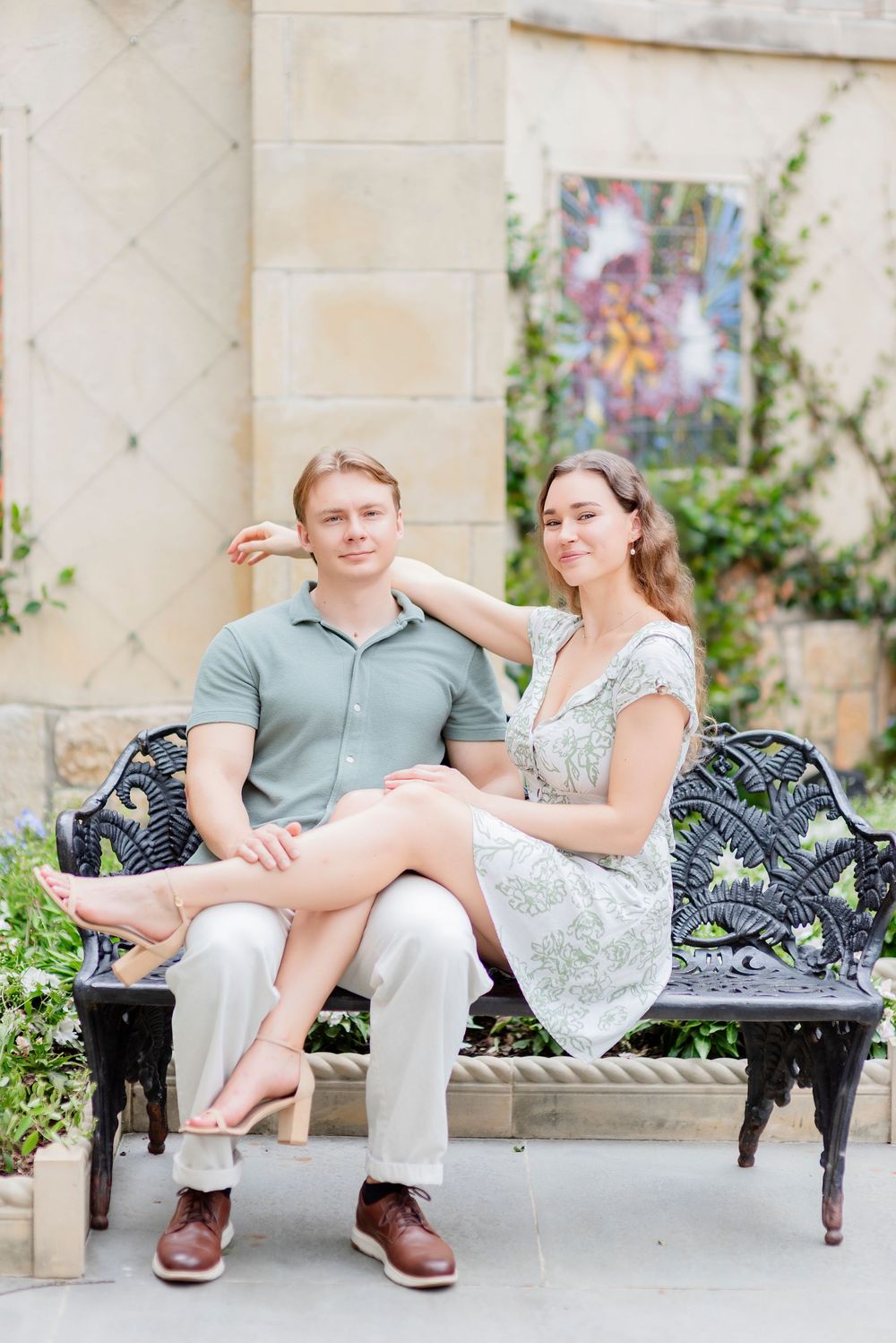 A couple relaxes on a decorative iron bench against a light stone wall.