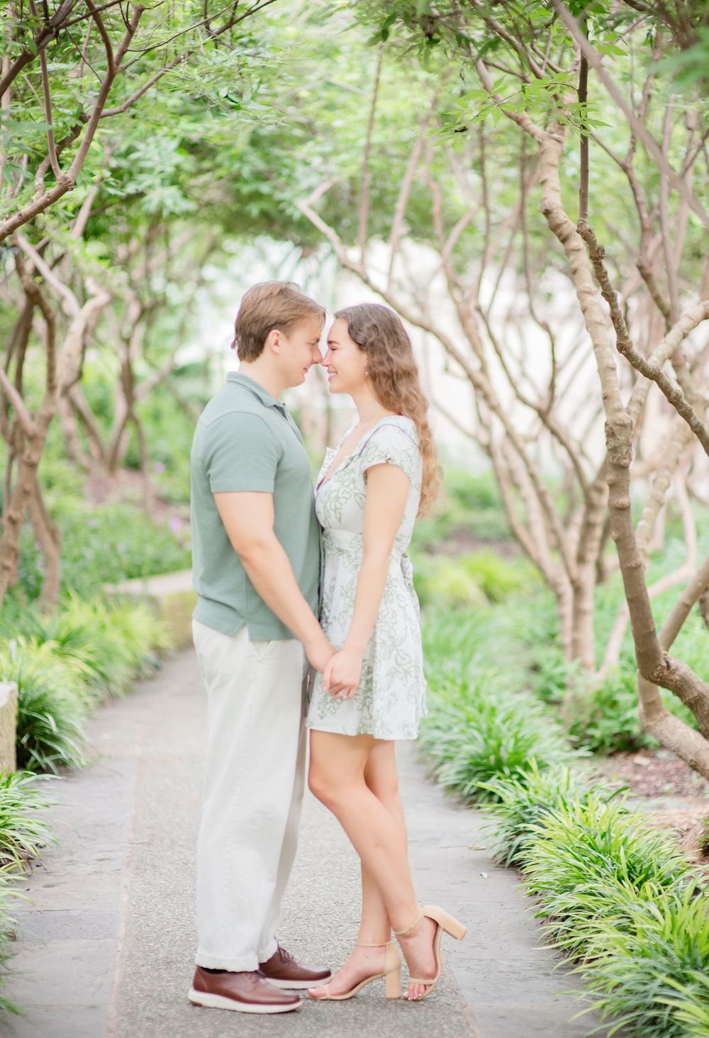 A romantic couple shares a tender moment on a garden path surrounded by lush greenery and trees.