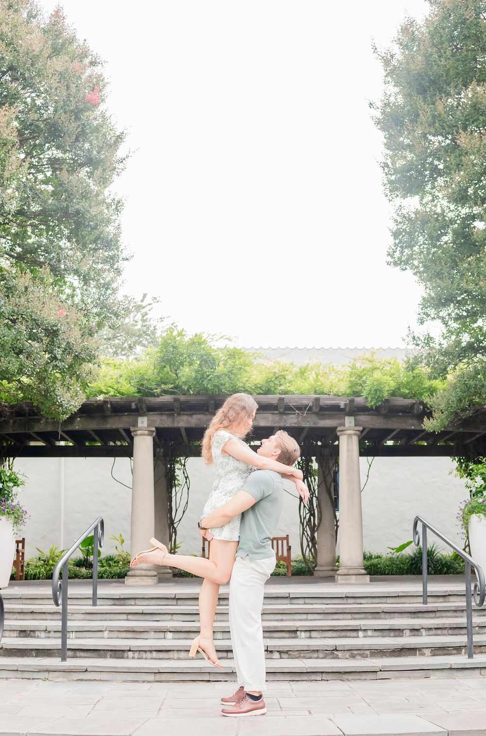 A playful couple embraces in front of a garden pergola with stone columns.