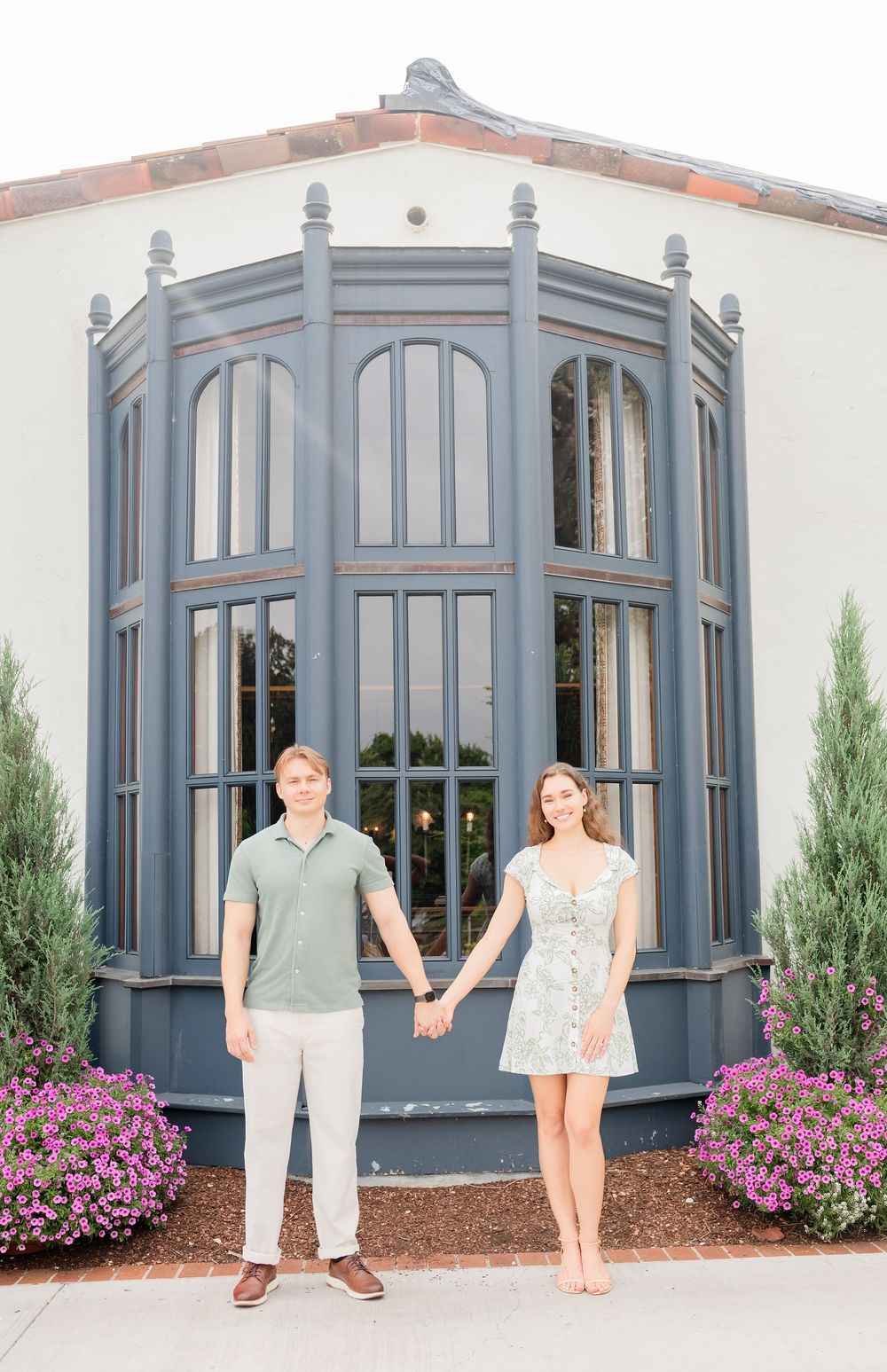A couple poses hand-in-hand in front of a beautiful grey conservatory with pink flowers and cypress trees.