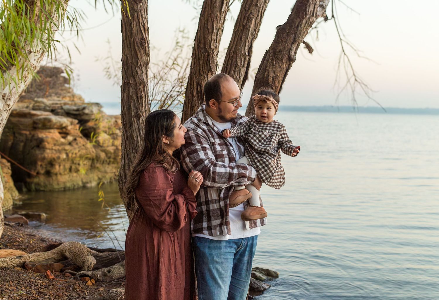 Couple in casual attire standing together near a scenic lake with rocky shoreline during golden hour.