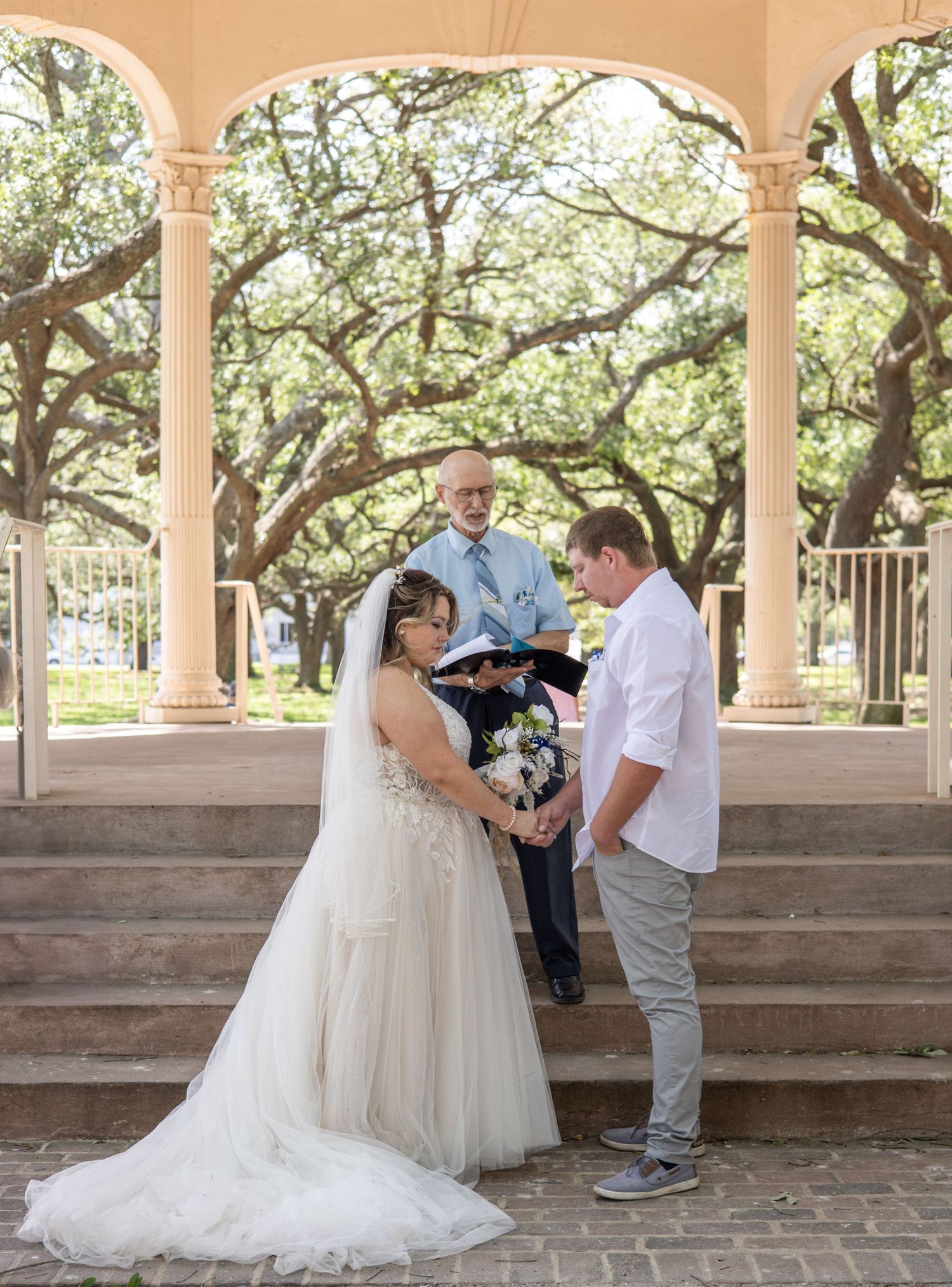 A wedding ceremony takes place under a beautiful archway with sprawling oak trees in the background.