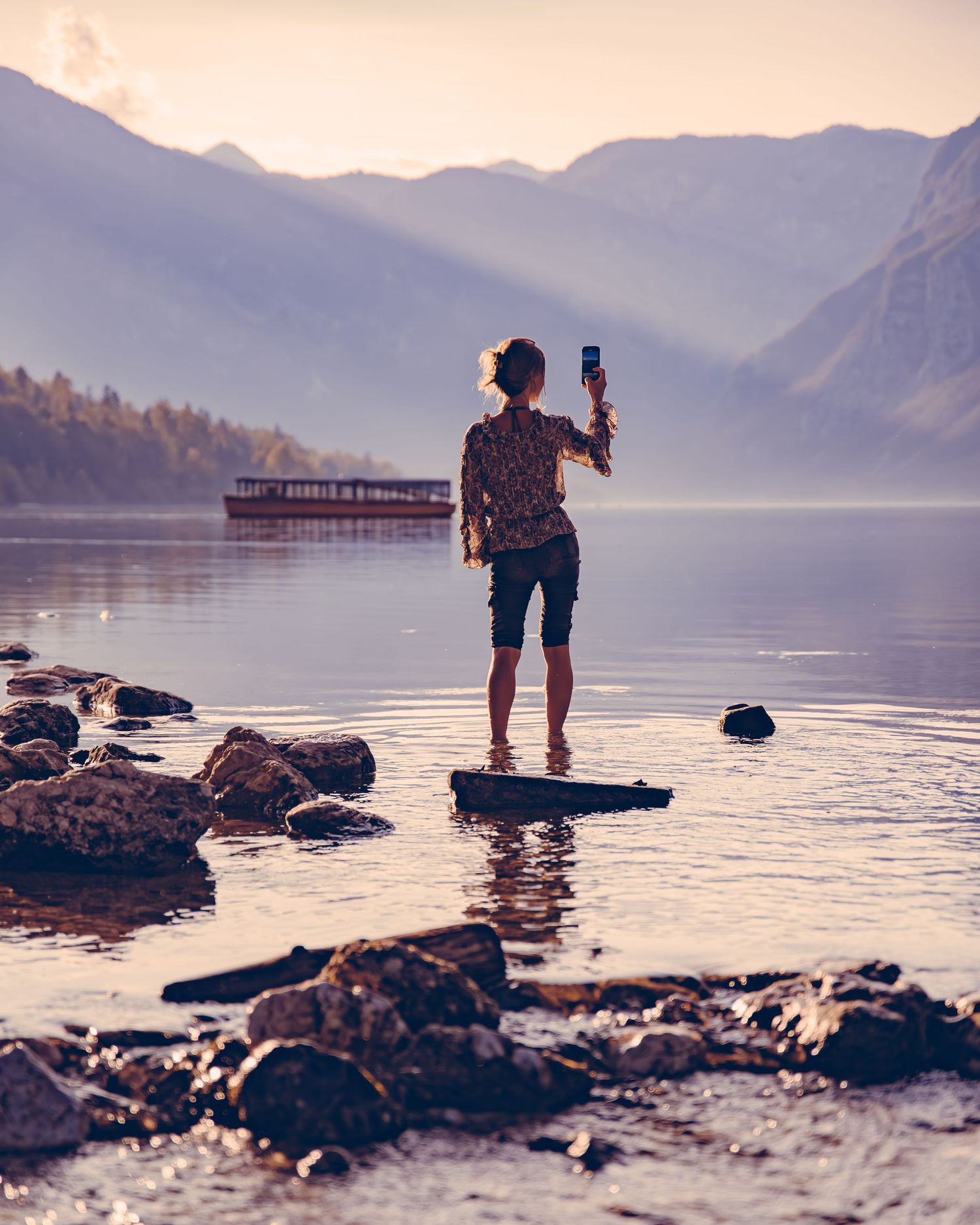 A silhouetted figure stands on rocks by a serene lake at sunset with mountains and a boat in the background.