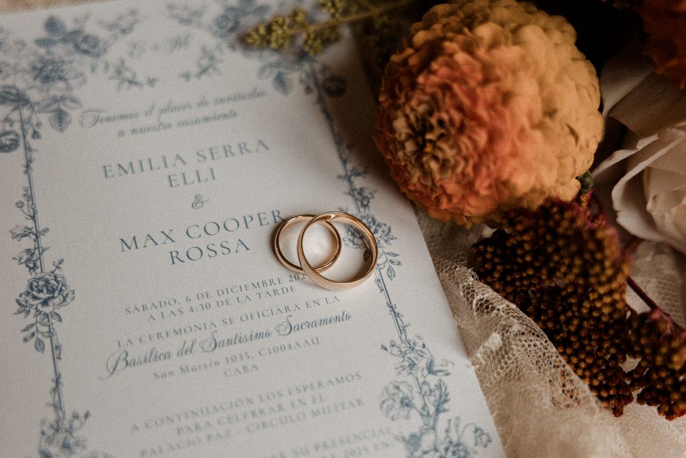 Wedding rings rest on elegant blue and white floral invitation cards with delicate cake pops in the background.