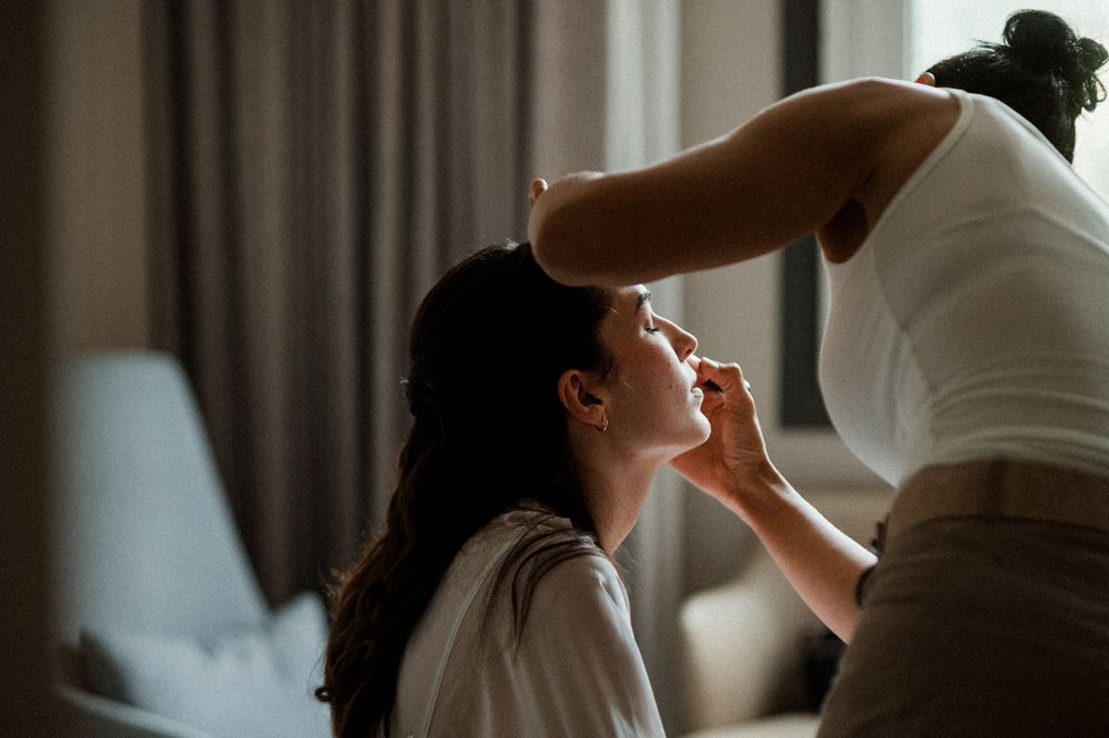 Makeup artist applies cosmetics to a client sitting in a chair by a window in soft natural lighting.